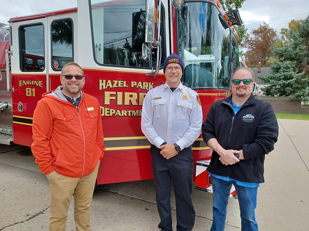 Three men smiling in front of a fire truck, labeled 'Hazel Park Fire Department, Engine 81.' The man in the center wears a fire chief uniform, while the others wear casual jackets. The background features trees and park buildings.