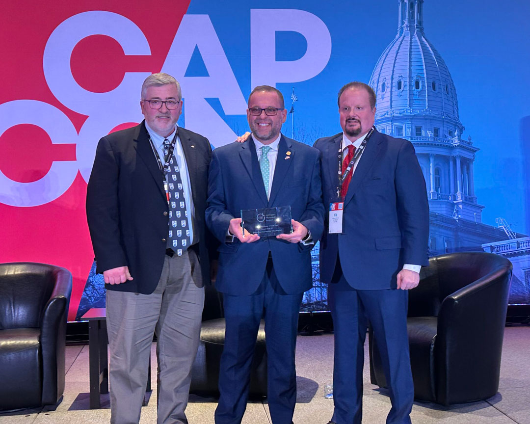 Three men posing together at a conference event. The center figure holds an award, dressed in a suit, while the other two wear blazers and ties. A backdrop featuring the words 'CAP CON' and a prominent building is visible.