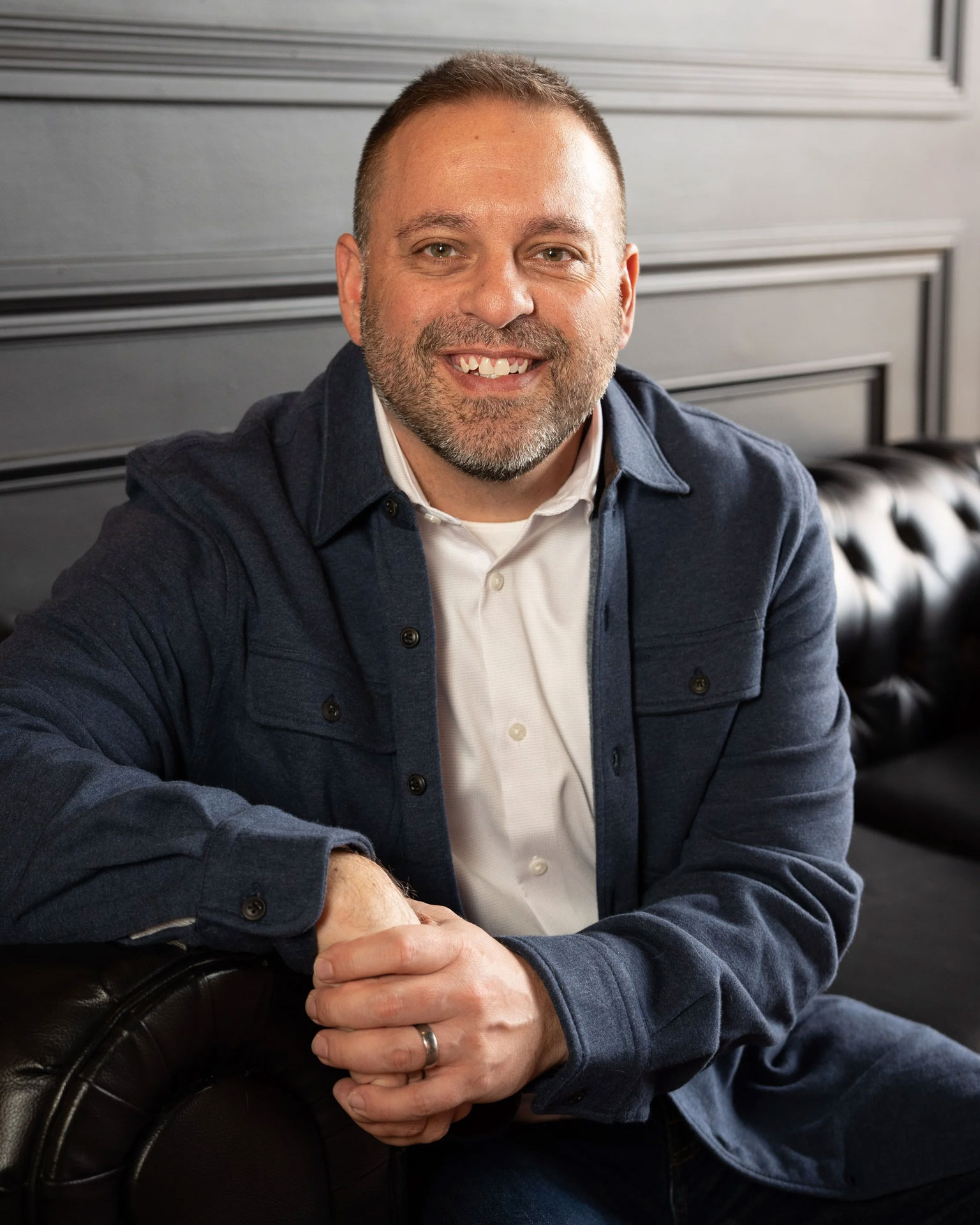 A man named McFall sitting comfortably on a black leather couch, wearing a dark blue shirt jacket over a white shirt. He is smiling and resting his right arm on his lap, with a modern interior backdrop featuring dark walls.