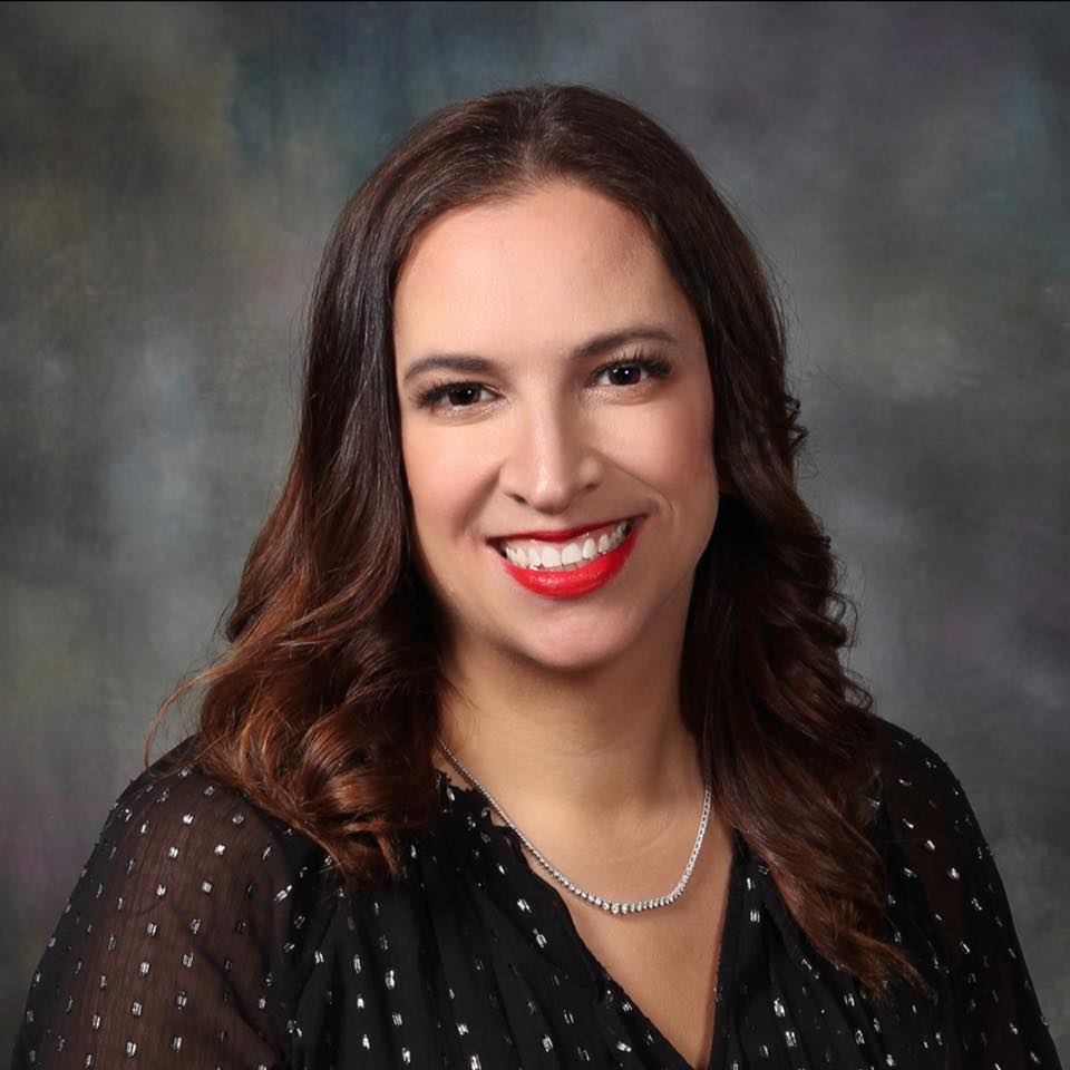 A portrait of Lori Barnwell with shoulder-length dark hair and a subtle wave. She is wearing a black blouse with polka dots and a silver necklace. Lori has a bright red lipstick smile and is set against a blurred gray background.