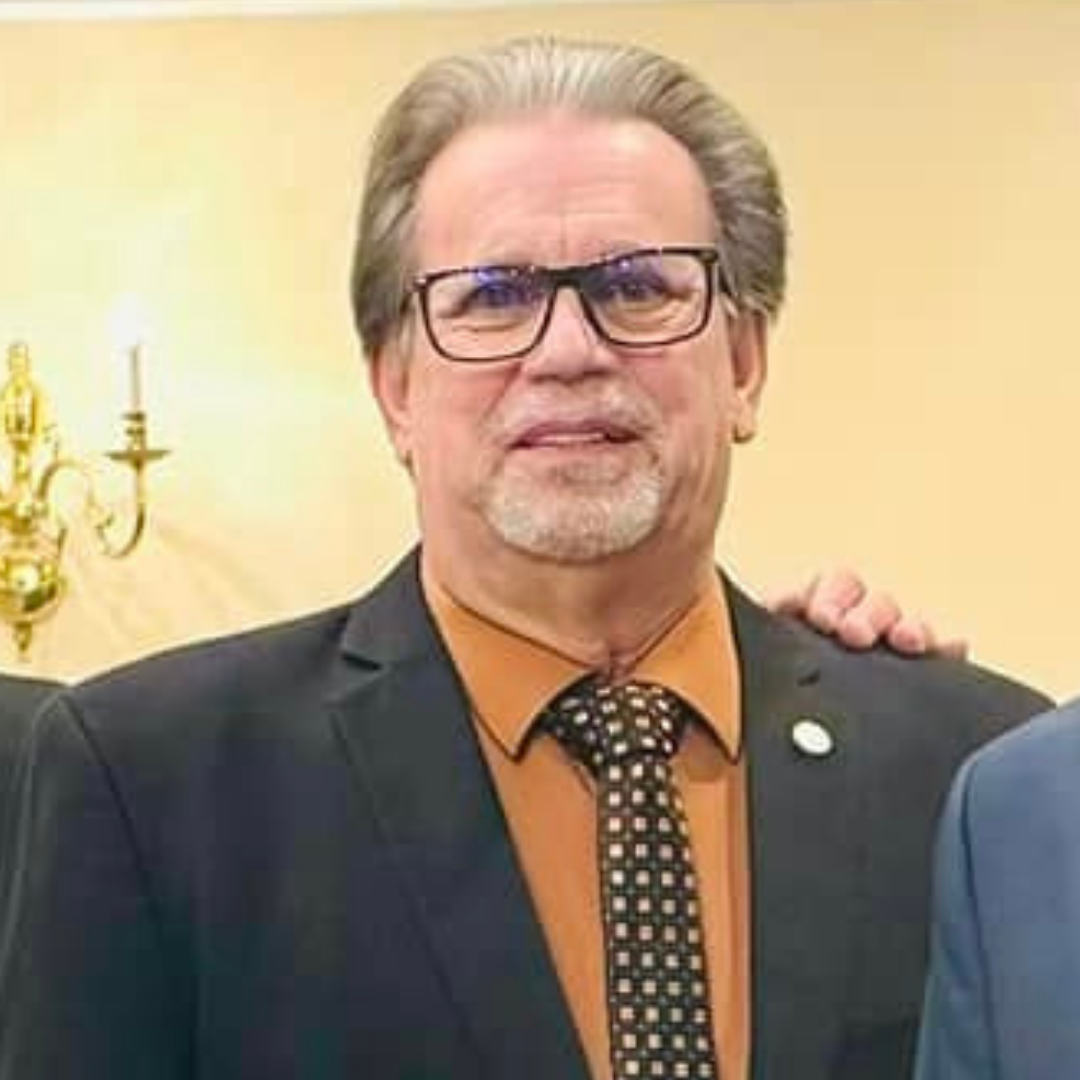 A middle-aged man with glasses and gray hair stands smiling. He wears a dark suit jacket over a brown shirt with a polka dot tie. In the background, a wall with a wall sconce and blurred figures are visible. The image suggests a formal or social gathering.