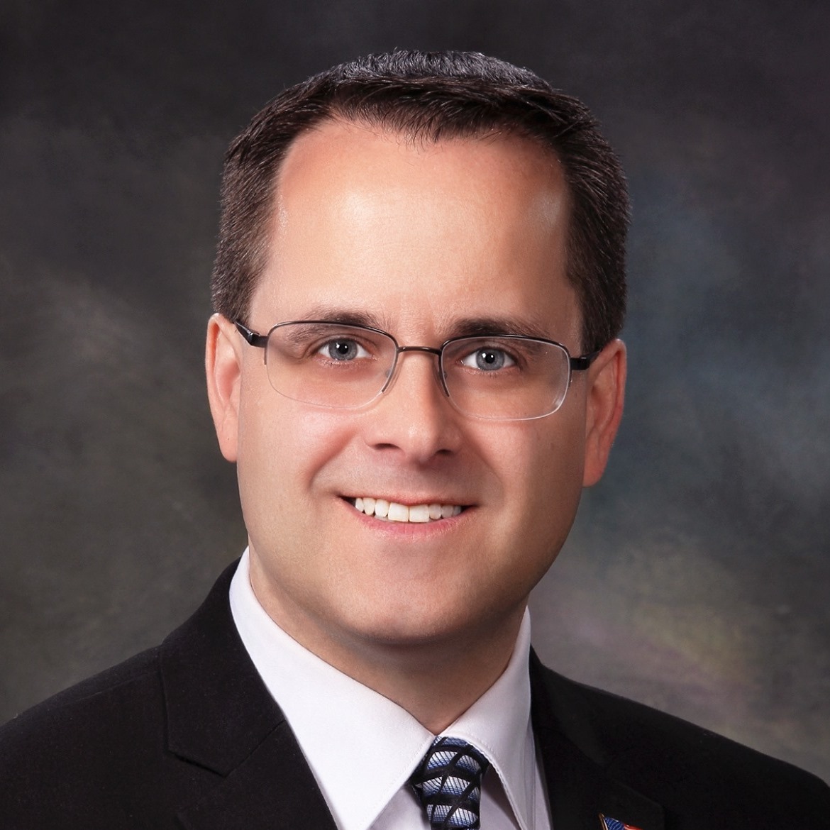 A headshot of Mindy Moore, a man wearing glasses and a dark suit with a white shirt and patterned tie. He has short, light brown hair and is smiling at the camera, set against a soft, blurred background.