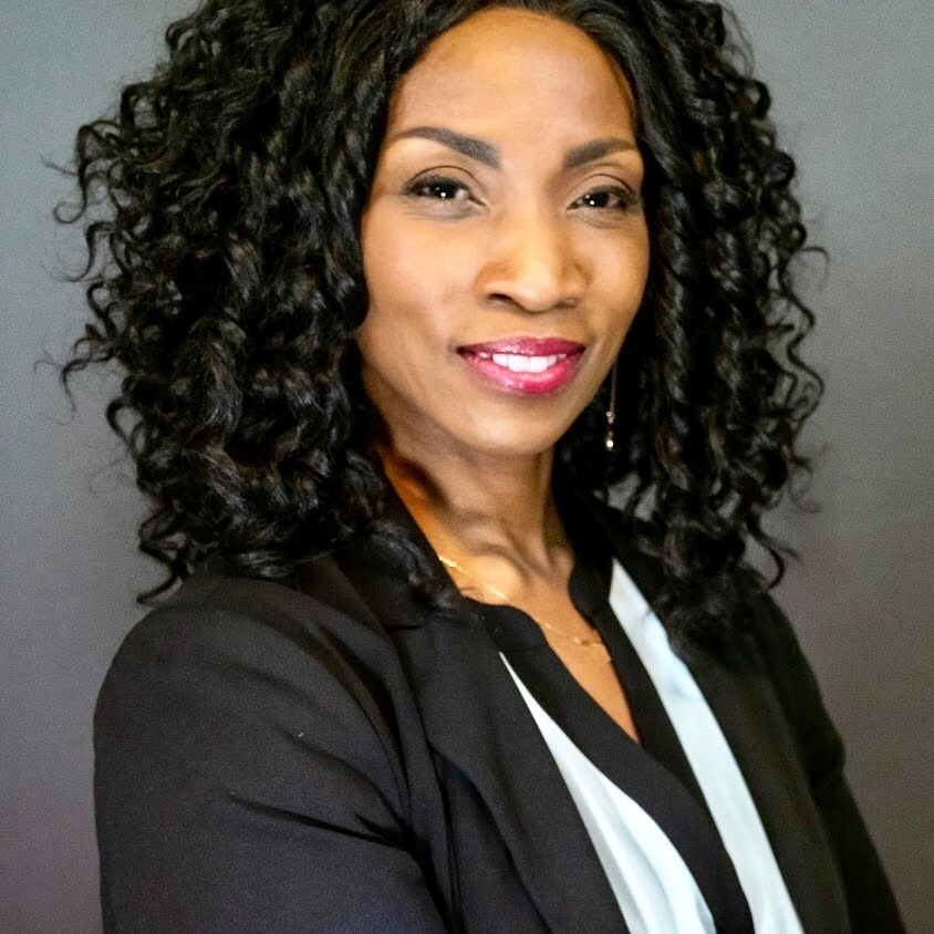 Mindy Moore poses for a professional headshot, wearing a black blazer over a white blouse. She has long curly hair and is smiling slightly, with a neutral background that enhances her appearance.