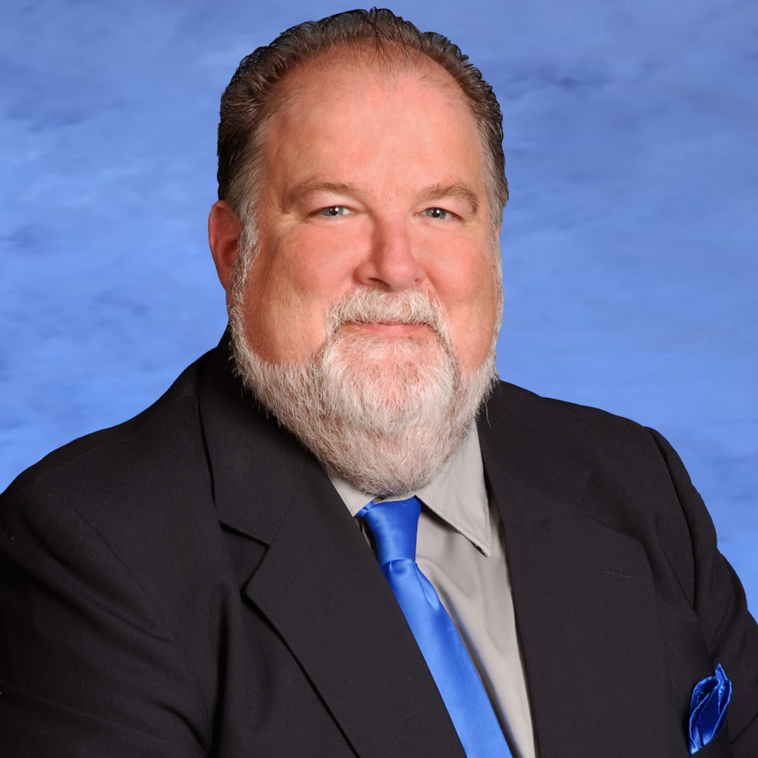 Portrait of William Mier, a man with a gray beard and mustache, wearing a black suit jacket over a gray shirt, and a blue tie. He is seated against a blue background, smiling at the camera.