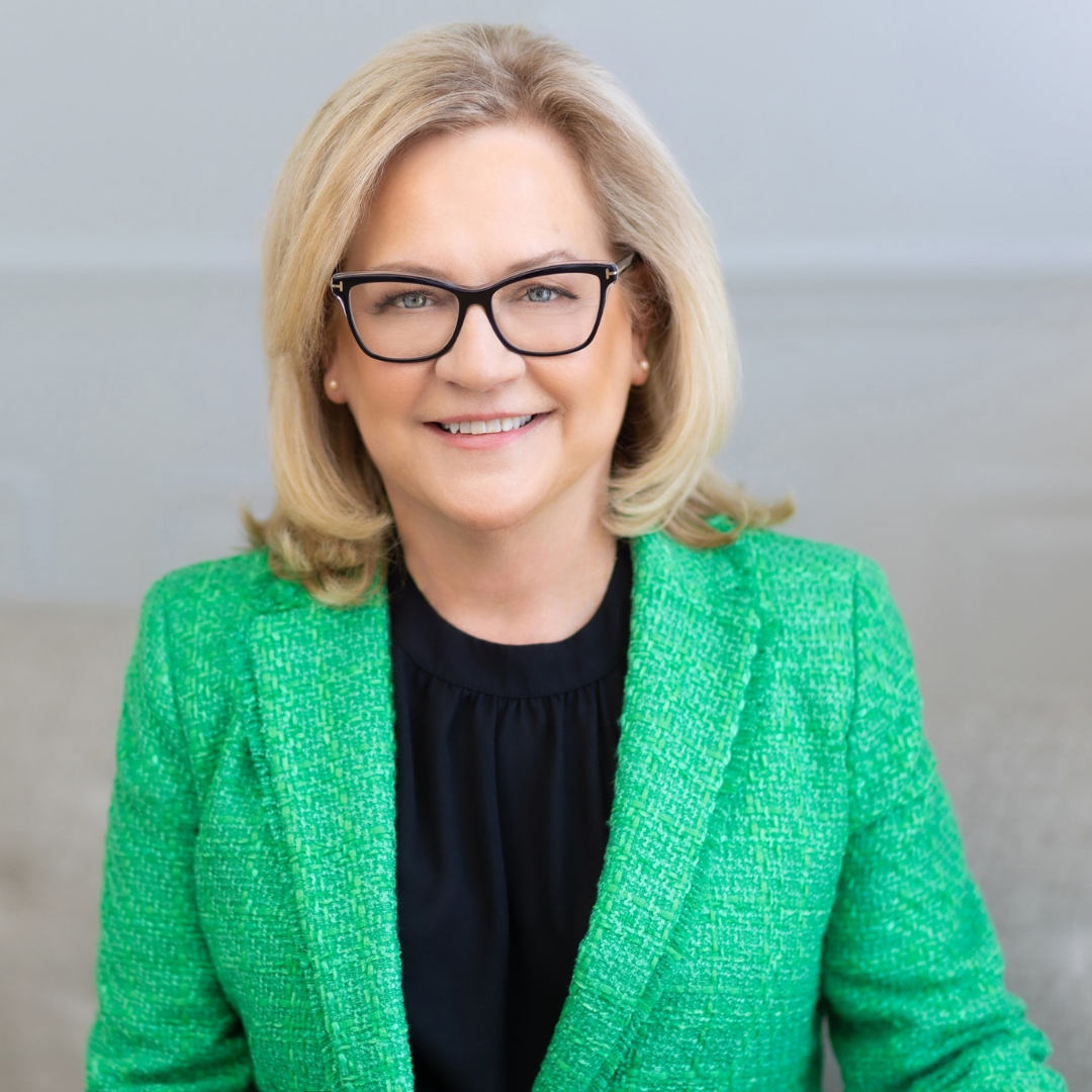 Mindy Moore is smiling and wearing glasses, dressed in a bright green blazer over a black top. She is seated in a well-lit environment, with a neutral background. Her light blonde hair is styled in loose waves.