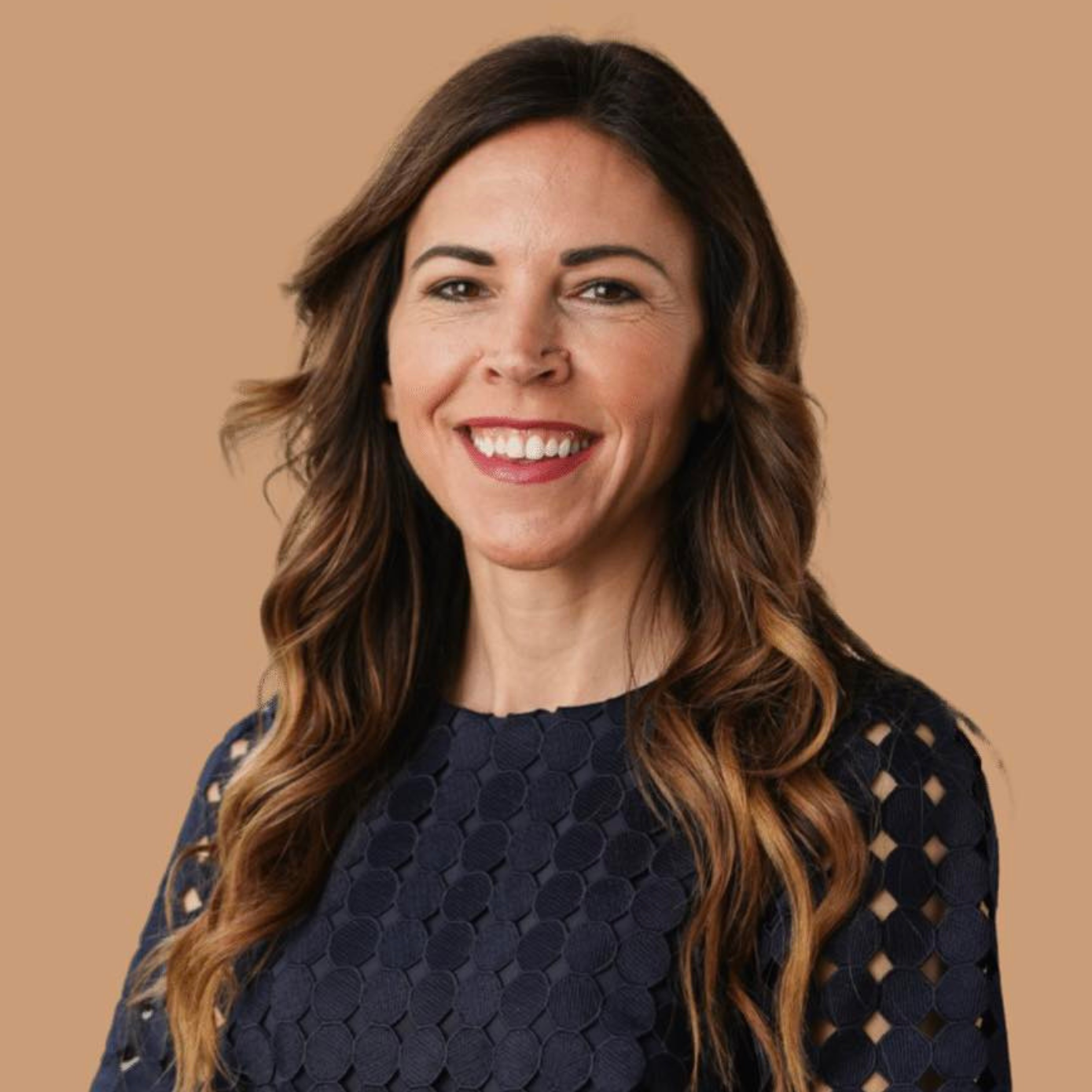 A woman with long, wavy brown hair smiles confidently at the camera. She wears a navy blue top with circular cutouts and has a neutral background. The image depicts a professional headshot.
