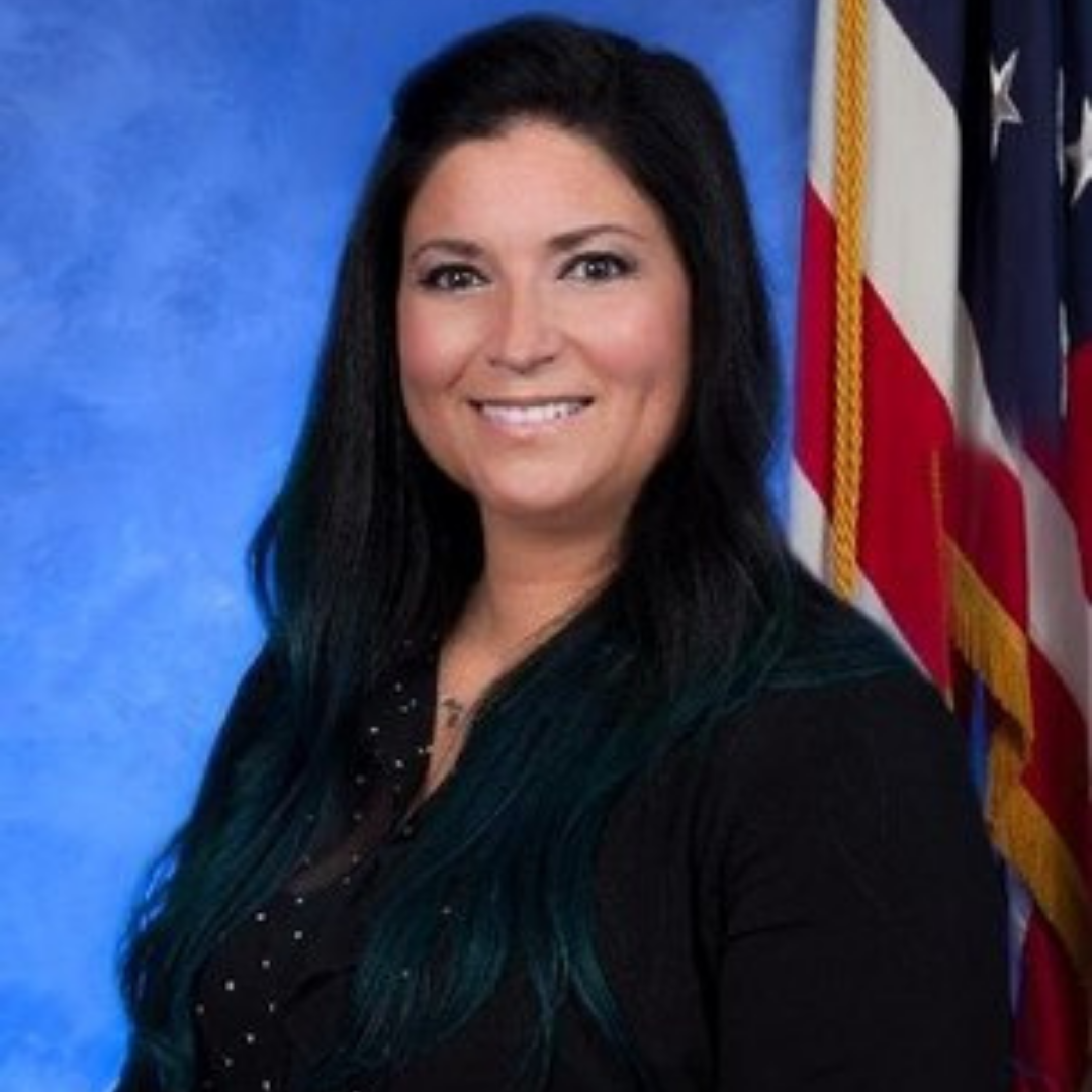 Alissa Sullivan, a woman with long black hair featuring teal highlights, smiles at the camera. She wears a black blouse with polka dots. In the background, there are American flags prominently displayed.