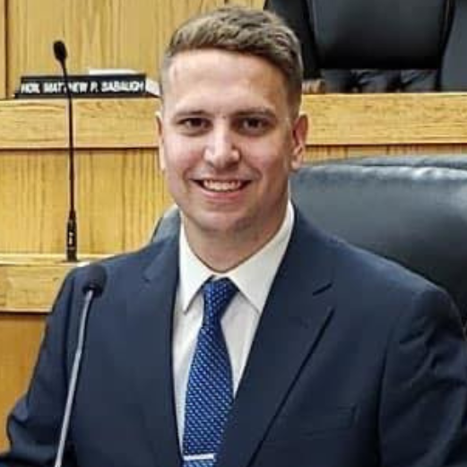 Aaron Delikta, a man with short hair, is seated at a conference table. He is wearing a navy suit with a white shirt and polka-dot tie, smiling at the camera. The background features wooden paneling and black leather chairs.