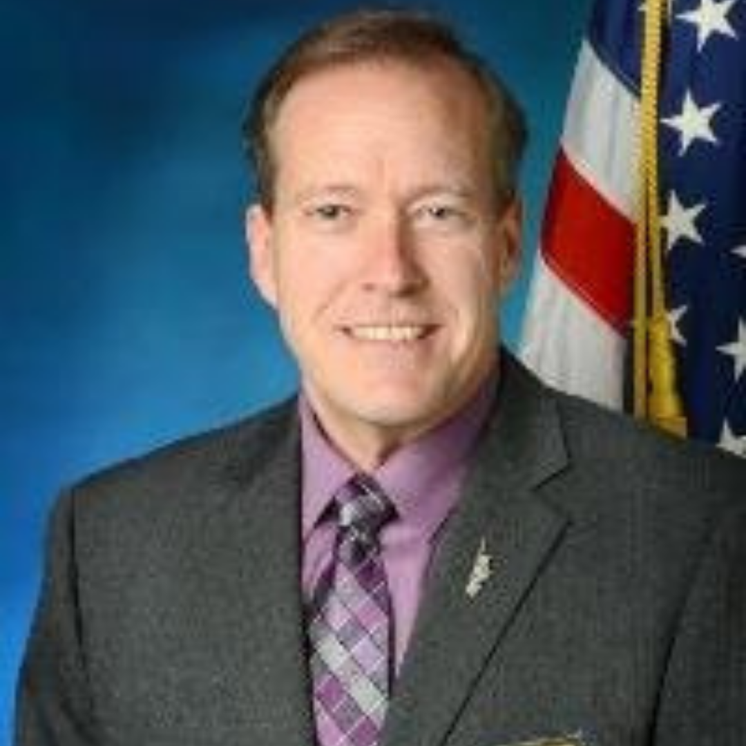 A man with short brown hair is smiling in front of a blue background. He is wearing a dark suit with a light purple shirt and a tie featuring a plaid pattern. There is a U.S. flag visible in the background.