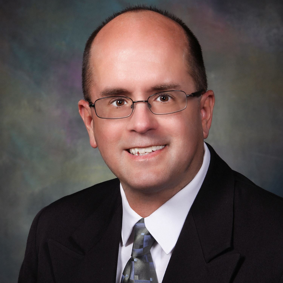 A professional headshot of Richard Moeller, featuring a smiling man with glasses, short brown hair, and a bald head. He wears a black suit and a light blue shirt with a patterned tie, set against a blurred green and gray background.
