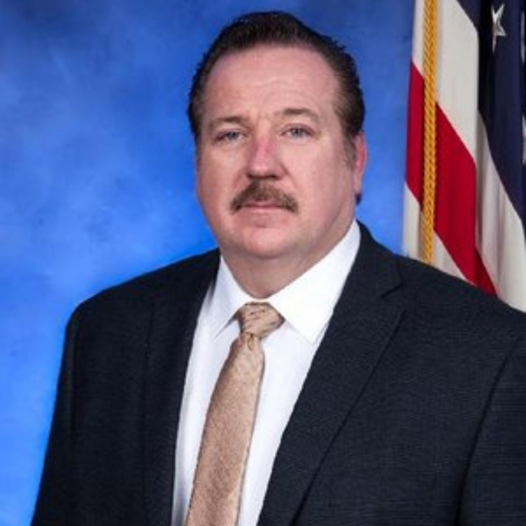 A man with a mustache wearing a dark suit and a white shirt, along with a beige tie, stands in front of a blue background. The image includes an American flag on the right side.
