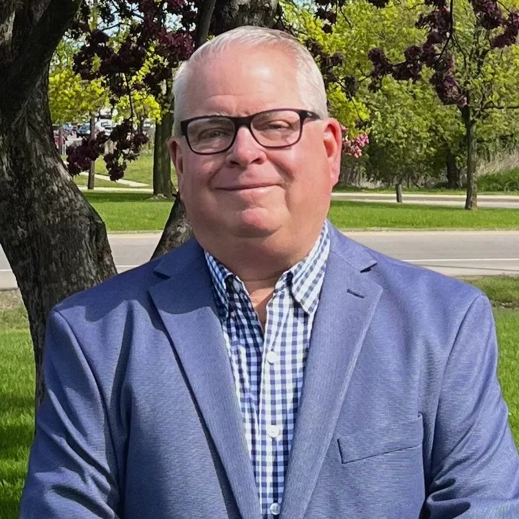 Dave Dwyer stands outdoors in front of a tree with green leaves. He is wearing a light blue blazer over a checkered shirt and glasses, with a slight smile, as he faces the camera.